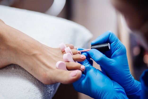 Thuis salon pedicure Voetverzorging en nagel Het proces van professionele pedicure Master in blauwe handschoenen brengt lichtroze gellak aan