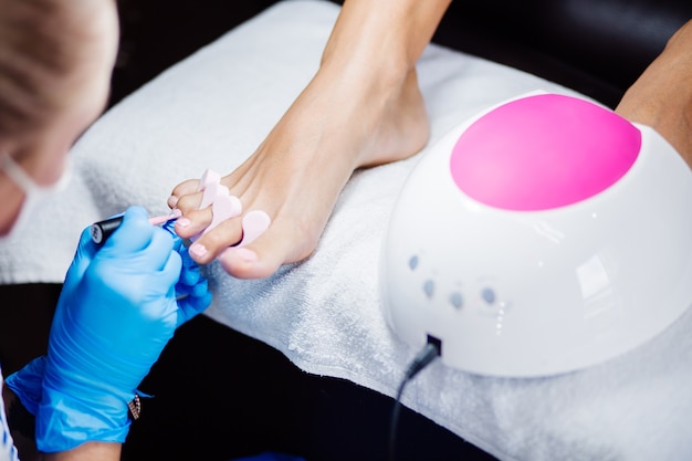 Thuis salon pedicure Voetverzorging en nagel Het proces van professionele pedicure Master in blauwe handschoenen brengt lichtroze gellak aan