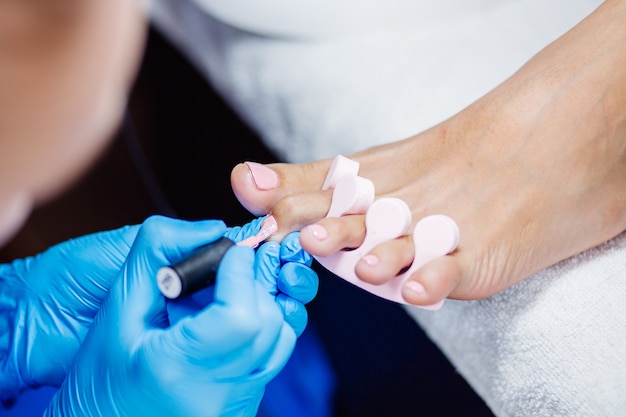 Thuis salon pedicure voetverzorging en nagel het proces van professionele pedicure master in blauwe handschoenen brengt lichtroze gellak aan