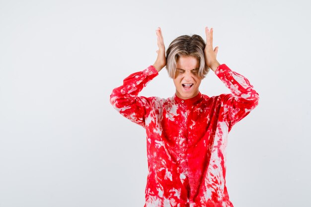 Tiener blonde man in oversized shirt met handen op het hoofd en op zoek pijnlijk, vooraanzicht.