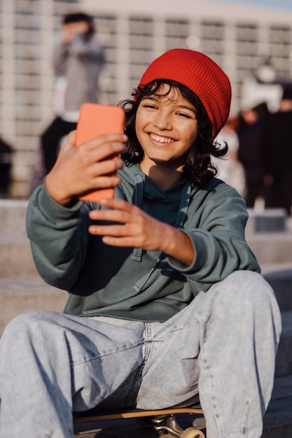 Gratis foto tiener buiten zittend op skateboard en selfie te nemen