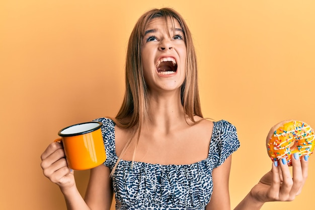 Gratis foto tiener kaukasisch meisje dat donut eet en koffie drinkt boos en boos schreeuwend gefrustreerd en woedend, schreeuwend van woede die omhoog kijkt.