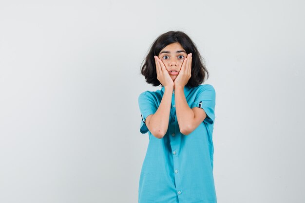 Tiener meisje in blauw shirt hand in hand op haar wangen en op zoek verrast