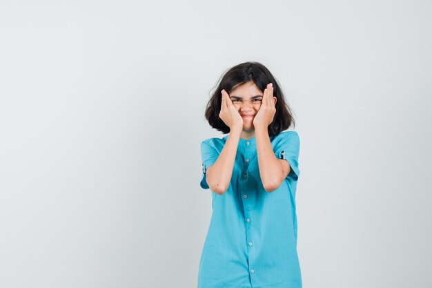 Tiener meisje omklemde gezicht met haar handen terwijl ze lacht in blauw shirt en er raar uitziet