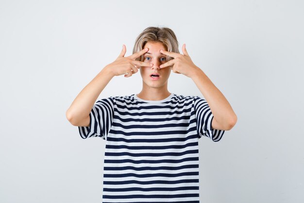 Tienerjongen die door vingers in t-shirt kijkt en geschokt kijkt, vooraanzicht.