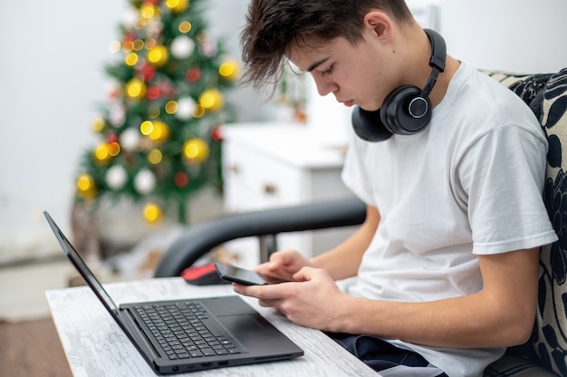 Tienerjongen gebruikt smartphone met koptelefoon, laptop op de knieën thuis. Kerstboom aan de muur. Serieus en gefocust gezicht