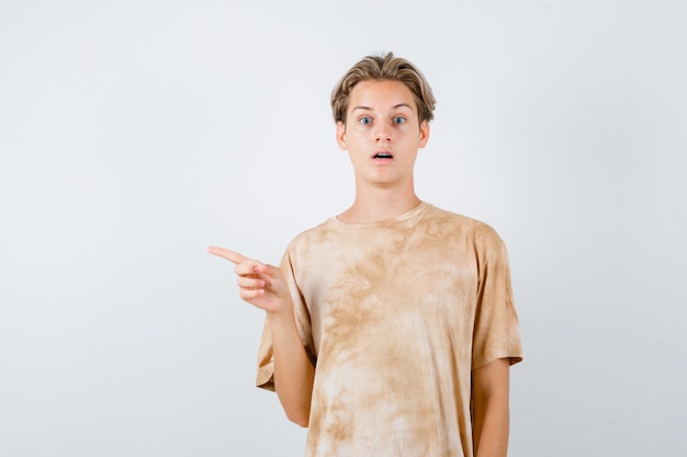 Tienerjongen in t-shirt die naar de linkerkant wijst, mond opent en geschokt kijkt, vooraanzicht.