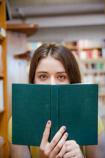 Gratis foto tienermeisje die gezicht behandelen met boek in bibliotheek