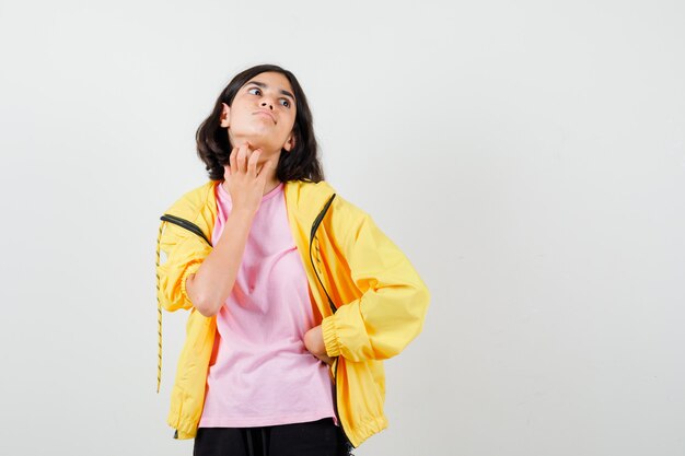 Tienermeisje in geel trainingspak, t-shirt dat nek krabt met hand en geschokt kijkt, vooraanzicht.