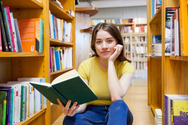 Tienermeisje met boek die camera tussen boekenkast bekijken