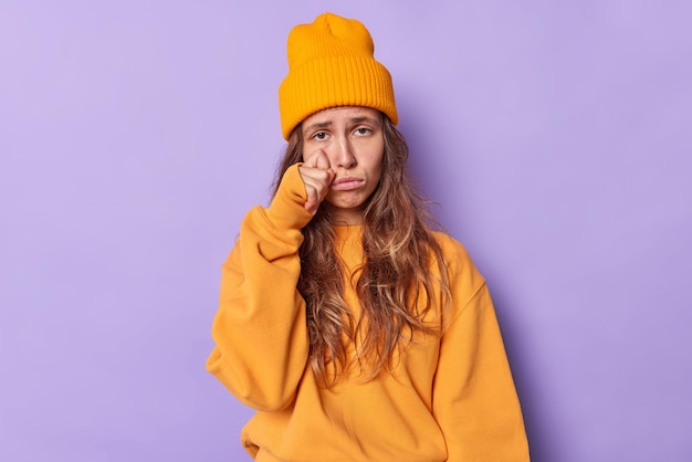 Tienermeisje wordt geconfronteerd met problemen snikt en veegt tranen staat erg overstuur draagt een casual oranje trui en hoed drukt negatieve emoties uit geïsoleerd op paars voelt zich diepbedroefd. Stress angst