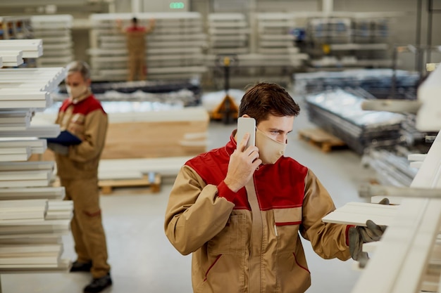 Timmerman met gezichtsmasker die aan de telefoon praat terwijl hij houten planken in een werkplaats onderzoekt