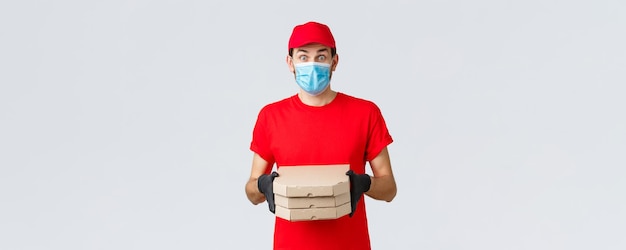 Toepassing voor voedselbezorging online boodschappen contactloos winkelen en covid19-concept Verrast koerier in rood uniform gezichtsmasker en handschoenen zien er onder de indruk uit en brengen klanten pizzadozen