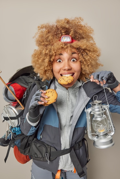 Toerisme en reizen concept verrast vrouw wandelaar voelt honger na lange afstand reizen eet koekjes houdt lantaarn draagt zware rugzak draagt jas en handschoenen poses tegen grijze achtergrond