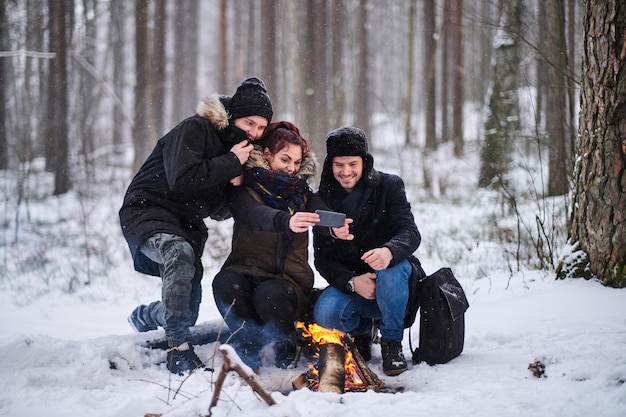 Toeristen zitten naast vreugdevuur selfie foto smartphone te nemen. Wandeling in het koude besneeuwde bos