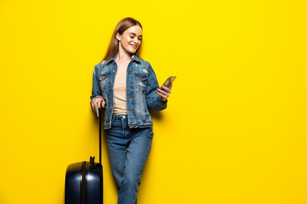 Toeristische vrouw met koffer in zomer casual kleding met telefoon geïsoleerd op gele muur