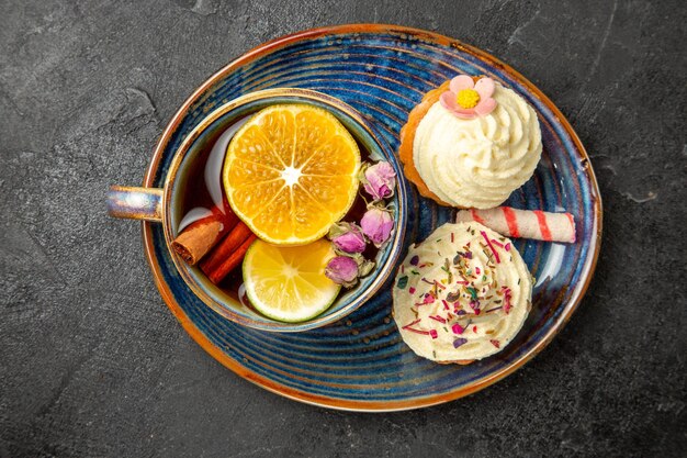 Top close-up een kopje thee met citroen twee smakelijke cupcakes met witte room snoep en een kopje met kruiden en citroen op de blauwe schotel op de zwarte tafel