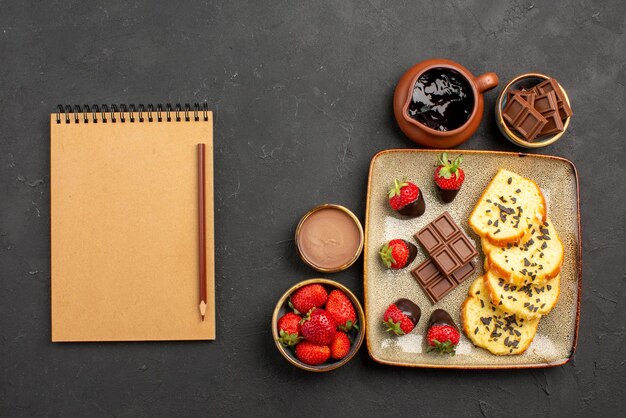 Top close-up weergave cake met chocolade notitieboekje en potlood naast de plaat van smakelijke cake met chocolade en aardbeien en aardbeien en chocolade crème in kommen