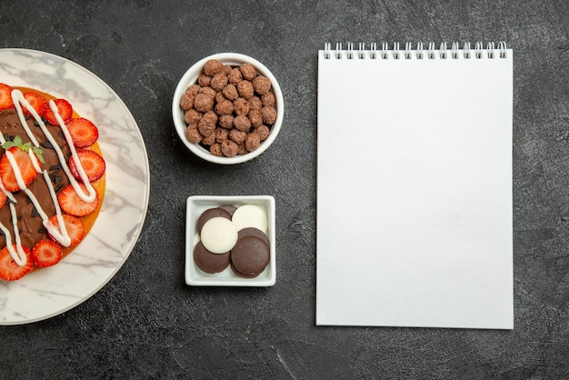 Top close-up weergave chocolade crème aardbeien kommen smakelijke chocolade en hazelnoten cake met bessen naast het witte notitieboekje