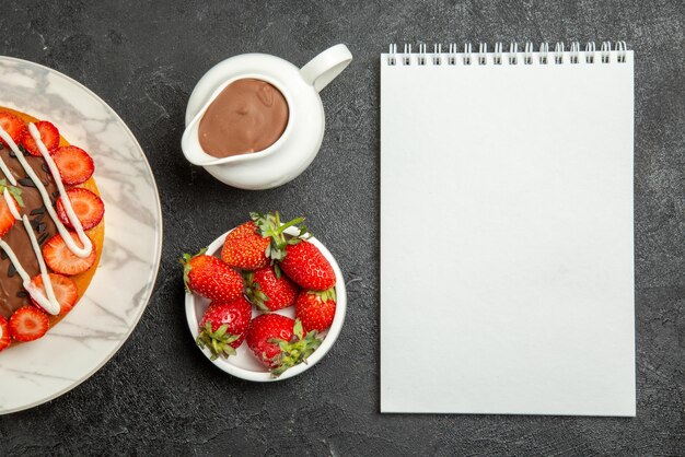 Top close-up weergave chocolade crème aardbeien smakelijke cake met bessen witte notebook kommen aardbeien en chocolade crème op tafel