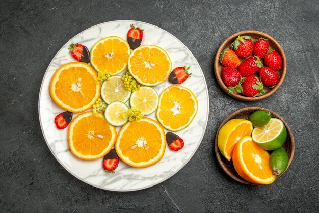 Top close-up weergave fruit op tafel witte plaat van oranje chocolade-bedekte aardbeien en citroen en bruine kommen van citrusvruchten en bessen op tafel