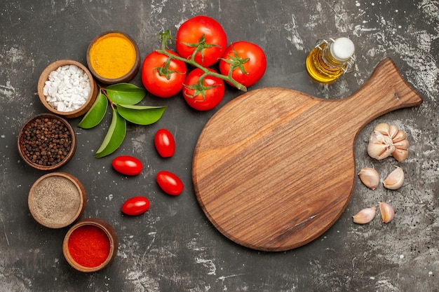 Top close-up weergave kruiden de snijplank naast de knoflook kommen van kleurrijke kruiden laat tomaten met steeltjes fles olie op de donkere tafel