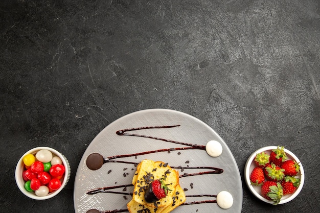 Top close-up weergave snoep grijze plaat van stukjes cake met chocoladesaus en aardbeien naast de kommen met snoep en aardbeien