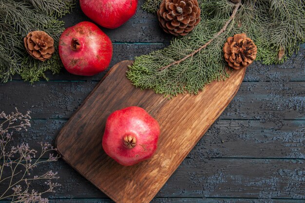 Top close-up weergave takken en granaatappels smakelijke granaatappel op keukenbord naast twee granaatappels en vuren takken met kegels op grijze tafel