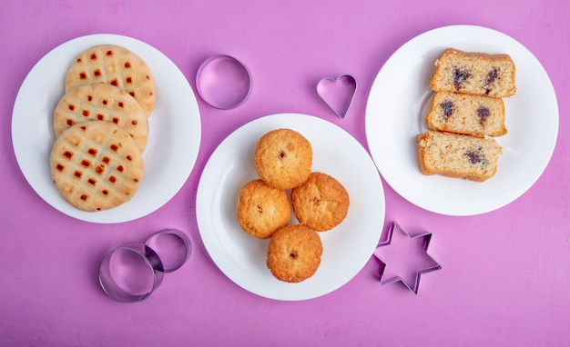 Topv uitzicht op koekjes en muffins op platen en cookie cutters op paarse achtergrond