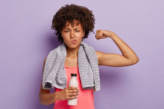 Training en sport concept. Gezonde atletische vrouw heft arm op, toont biceps, draagt casual roze top, houdt fles water vast, heeft handdoek op nek voor het afvegen van zweet, oefeningen binnen