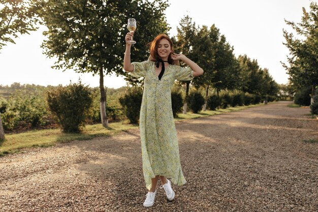 Trendy dame met rood kapsel in hoed, lange gele jurk en witte sneakers glimlachend en met glas met wijn op de achtergrond van de weg