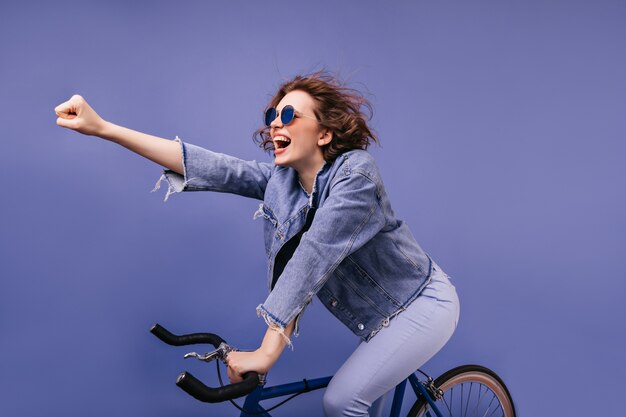 Trendy dame zittend op de fiets lachen en zwaaiende hand. Portret van schattige blanke vrouwelijke fietser.