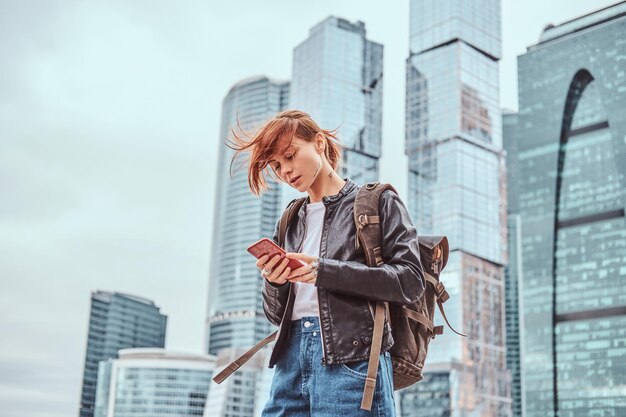 Trendy gekleed roodharige student meisje met tatoeages op haar gezicht met behulp van een smartphone voor wolkenkrabbers in Moskou stad op bewolkte ochtend.