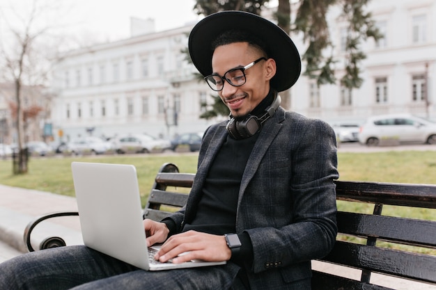 Trendy internationale student die met laptop in park werkt. Glimlachende Afrikaanse man buiten koelen met computer.