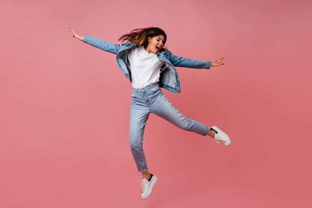 Trendy jonge vrouw springen op roze achtergrond. Volle lengte weergave van onbezorgd vrouwelijk model in denim outfit.