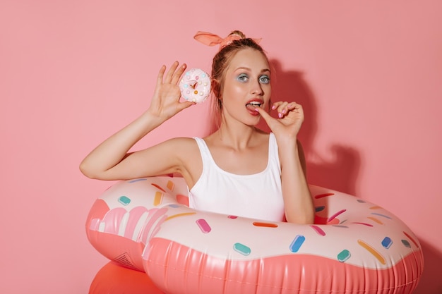 Gratis foto trendy meisje met moderne make-up in lichte stijlvolle outfit poseren met donut en roze zwemringen op geïsoleerde achtergrond