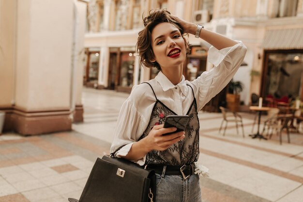 Trendy meisje met rode lippen en kort haar in shirt met zwart kant dat camera in de stad onderzoekt Vrouw met handtas en telefoon op straat