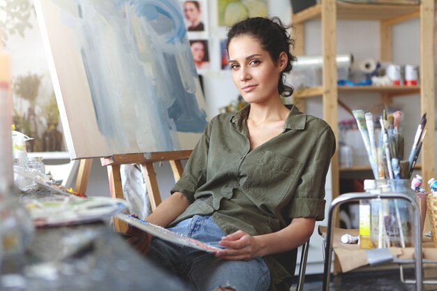 Trendy ogende positieve getalenteerde jonge blanke vrouw kunstenaar ontspannen op stoel naast ezel in werkplaats nadat ze klaar was met schilderen. Beroep, creatieve, moderne kunst, beroep en beroep
