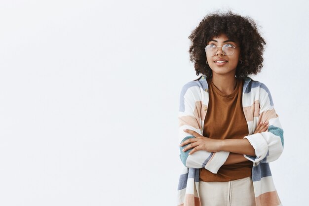 trendy stedelijke Afro-Amerikaanse vrouwelijke werknemer in transparante trendy bril en outfit hand in hand gekruist op de borst en starend naar de linkerbovenhoek met een dromerige schattige glimlach