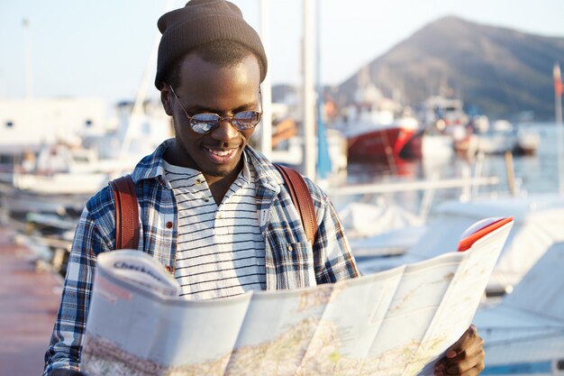 Trendy uitziende Afro-Amerikaanse toerist met rugzak in hoed en zonnebril die richtingen bestudeert met behulp van stadsgids terwijl hij bezienswaardigheden en monumenten van de badplaats verkent