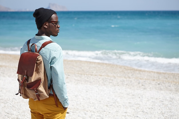 Trendy uitziende jonge Afro-Amerikaanse reiziger die een knapzak draagt die naar de kalme azuurblauwe zee voor hem kijkt, met een bedachtzame dromerige uitdrukking, staande op een kiezelstrand