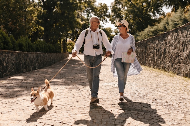 Trendy vrouw in hoed en blauwe blouse wandelen en zoek grijze harige man in wit shirt met lange mouwen met camera en corgi in park.