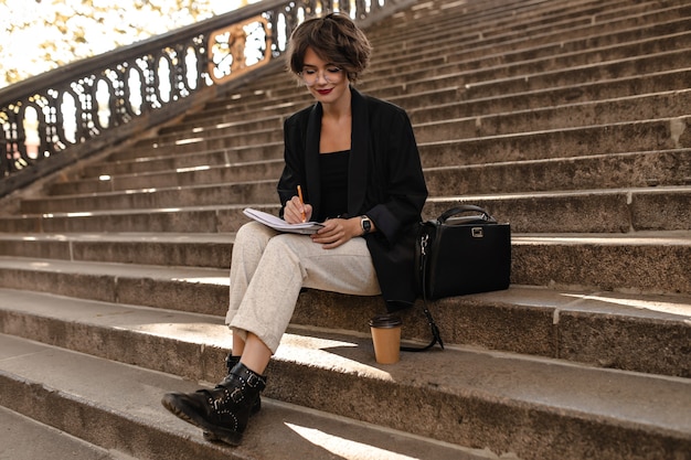 Trendy vrouw in lichte broek, zwarte jas en laarzen zit buiten op de trap. Kortharige dame in glazen buiten schrijven.