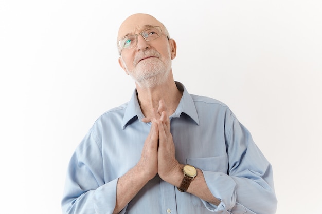 Treurige oude volwassen man in bril en blauw shirt met hoopvolle gezichtsuitdrukking, handen tegen elkaar gedrukt in gebed, in de hoop op het beste terwijl hij moeilijkheden, stress of problemen onder ogen ziet