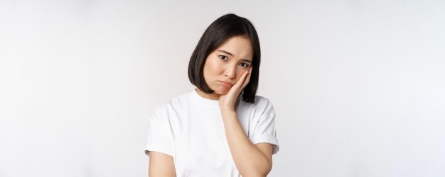 Triest aziatisch meisje dat boos en eenzaam mokkend en fronsend tegen een witte achtergrond in een casual t-shirt staat
