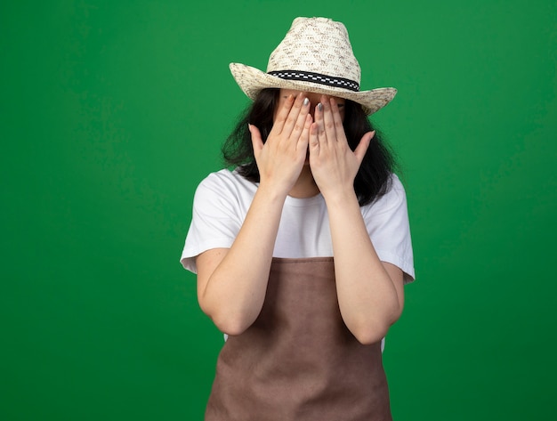 Triest jonge brunette vrouwelijke tuinman in optische bril en uniform dragen tuinieren hoed legt handen op gezicht geïsoleerd op groene muur