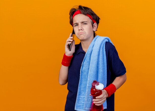 Triest jonge knappe sportieve jongen met handdoek op schouder, waterfles vasthouden en praten over de telefoon