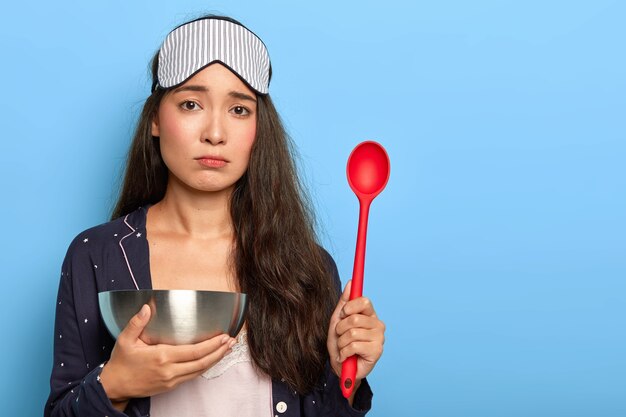 Triest mooie vrouw in nachtpak en slaapmasker, staat vroeg op voor het maken van ontbijt, kookt in de keuken, houdt kom met lepel vast, is in een lage geest, staat binnen