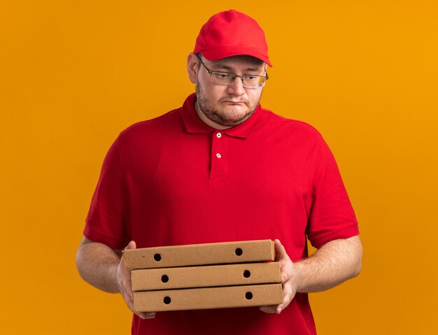triest overgewicht jonge bezorger in optische bril met pizzadozen naar beneden kijken geïsoleerd op oranje muur met kopieerruimte