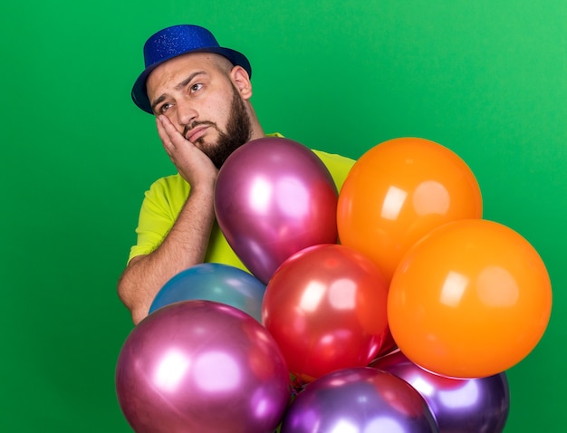 Triest uitziende jonge man met een feestmuts die achter ballonnen staat, bedekte wang met hand